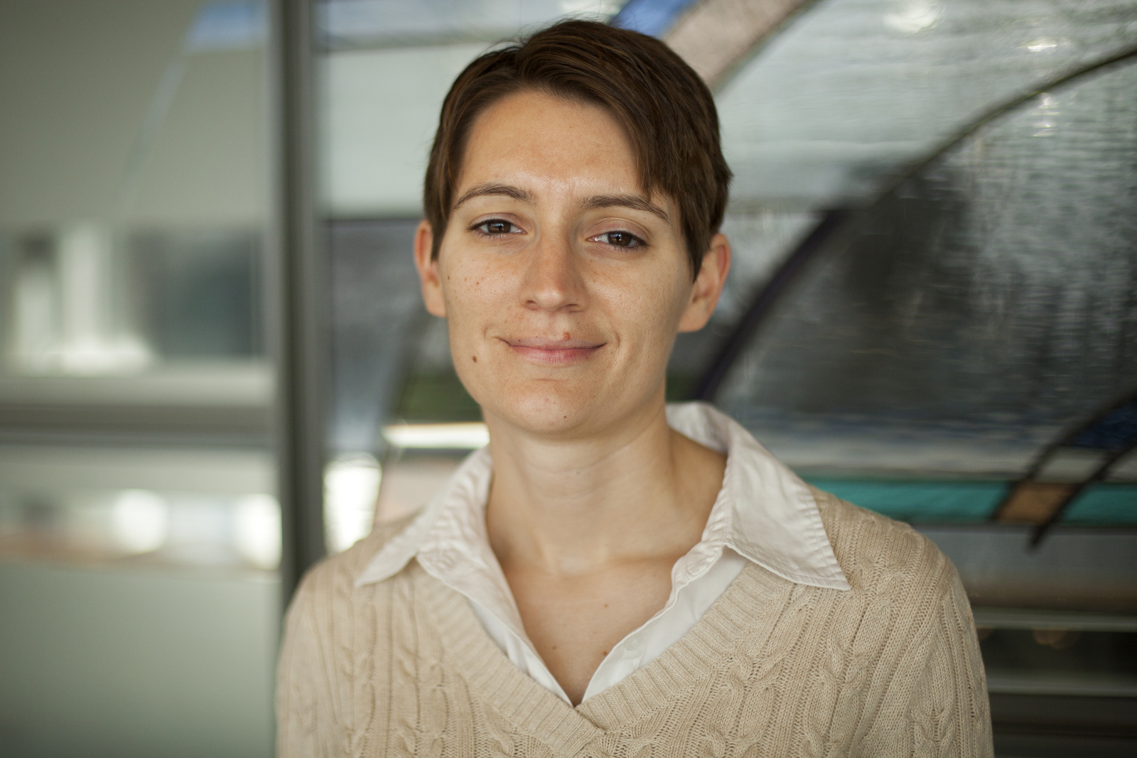 Ann Jeffers, story contributor, wearing a beige sweater on top of a white button-up shirt and standing in front of a glass background.