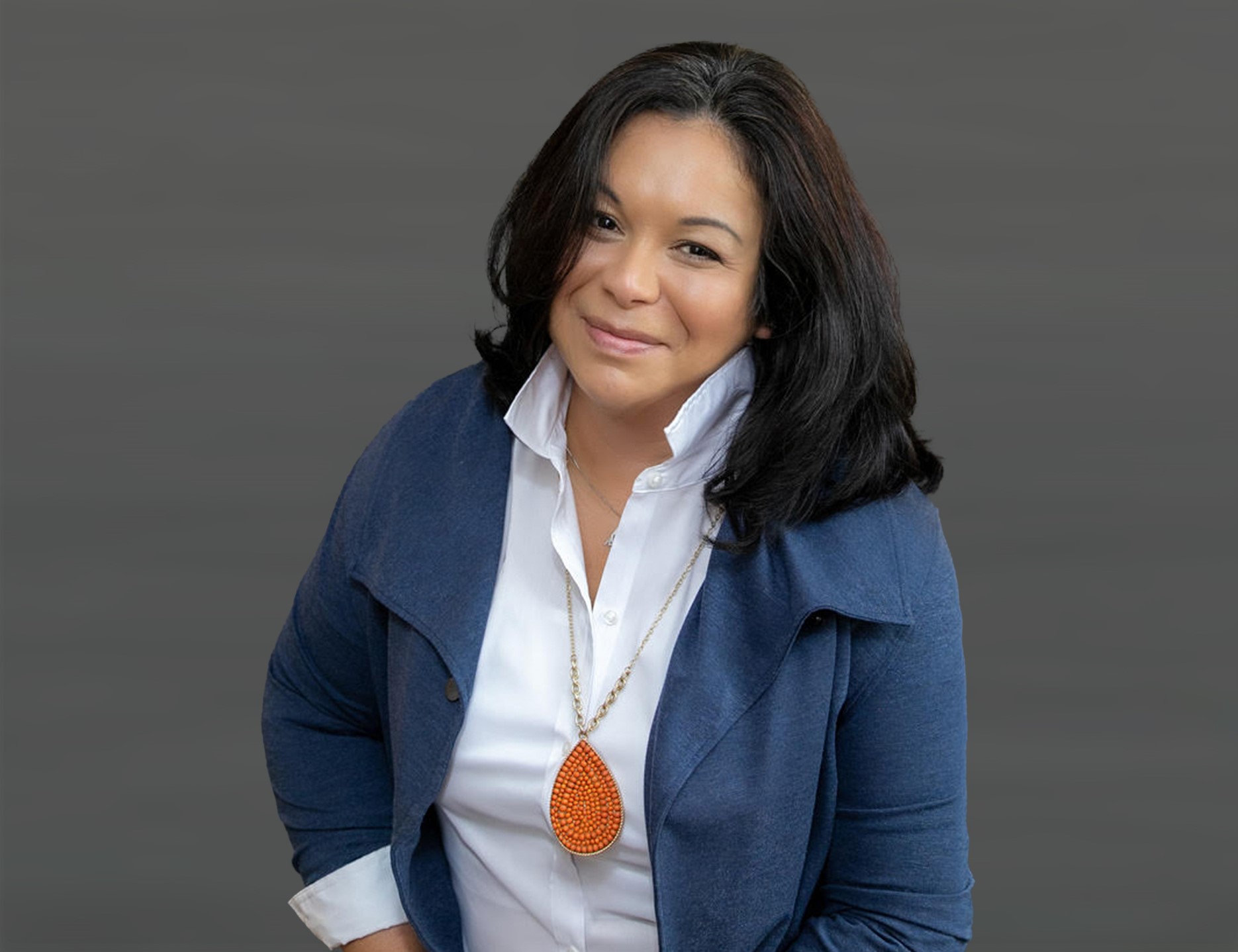 Annette Whittenberger, a story contributor, wearing a white collared shirt under a dark blue jacket and an orange teardrop-shaped pendant necklace. She's standing in front of a neutral background.