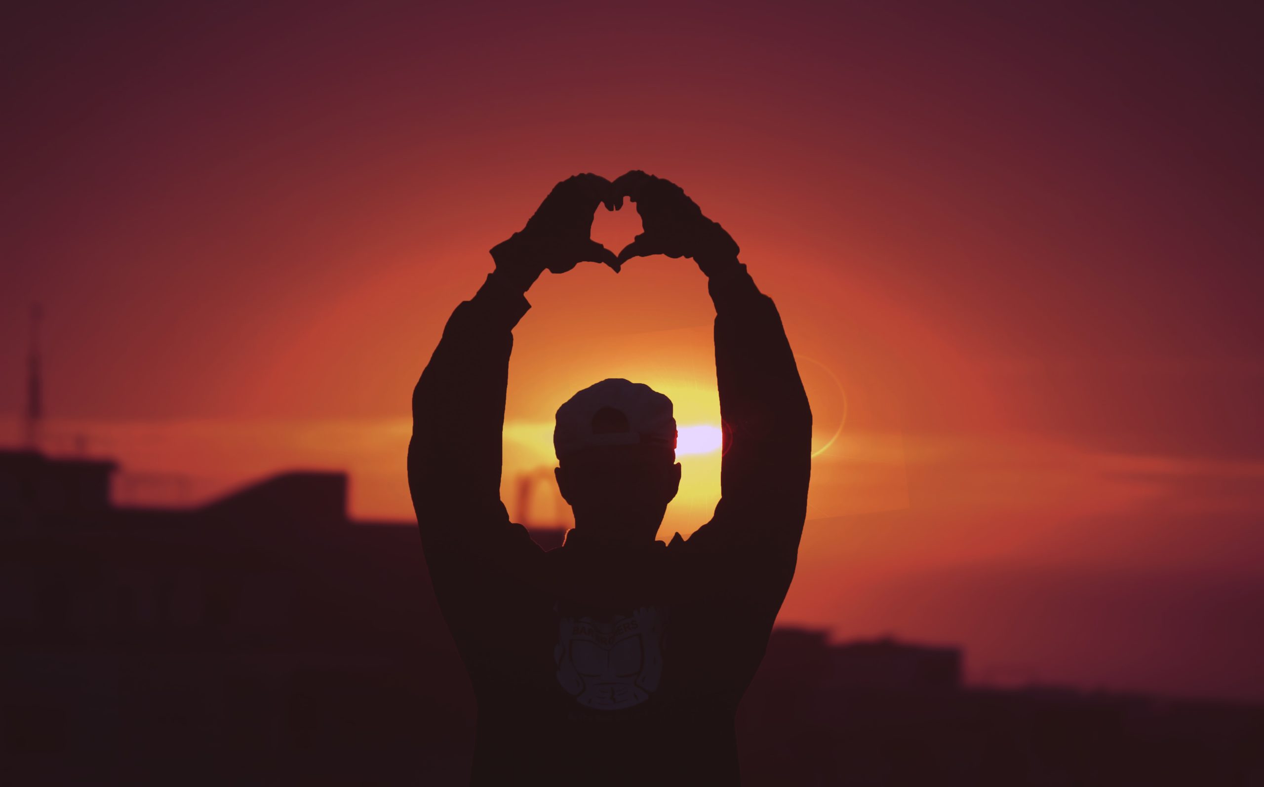 Silhouette of man making heart-shaped hand gesture at sunset.