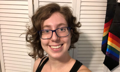 Erica Rae, contributor, wearing glasses and black tank top, in front of a white closet door from which hang two black ties with rainbows on them.