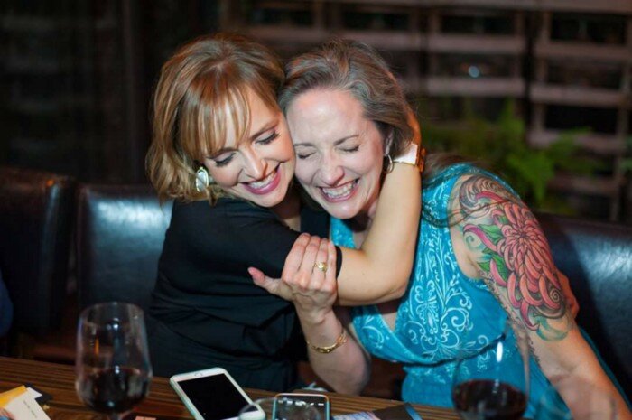 Two laughing friends hugging at a restaurant table. 