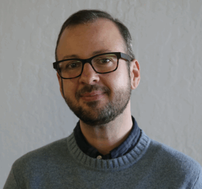Jules Plumadore, contributor, in dark-framed glasses and a beard, wearing a gray-blue wool sweater over a blue plaid button-down shirt, standing in front of a neutral background