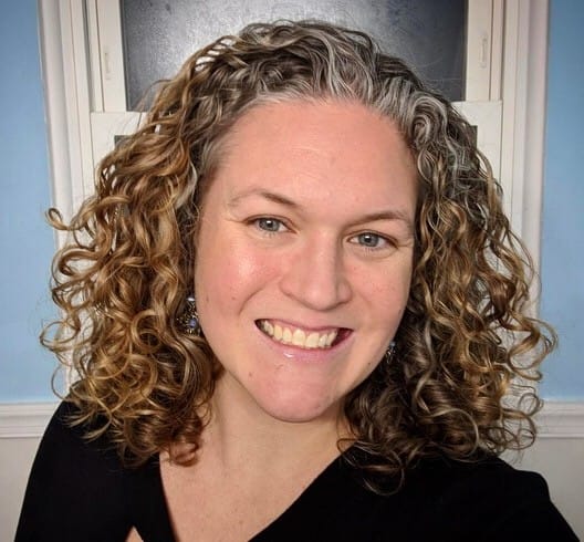 A smiling headshot of Laura Riordan, contributor; she has shoulder-length curly brown hair and is wearing a black V-neck top