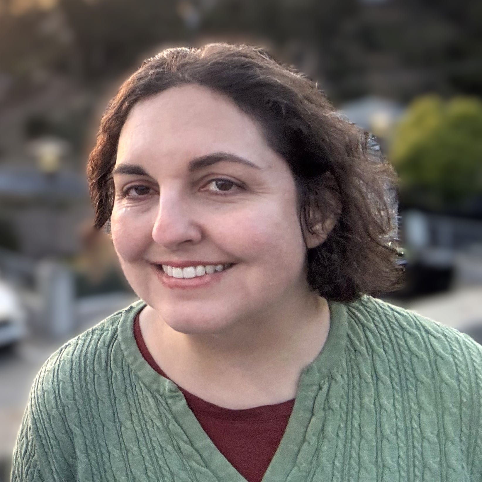 Rebecca, story contributor, has a wavy brown bob and is smiling at the camera; she is wearing a mossy green cardigan over a brown top. The background of the photo is blurred.