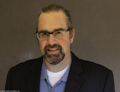Tim Dreby, contributor, wearing a blue button-down shirt under a dark blazer, glasses, and a goatee, in front of a brown backdrop.