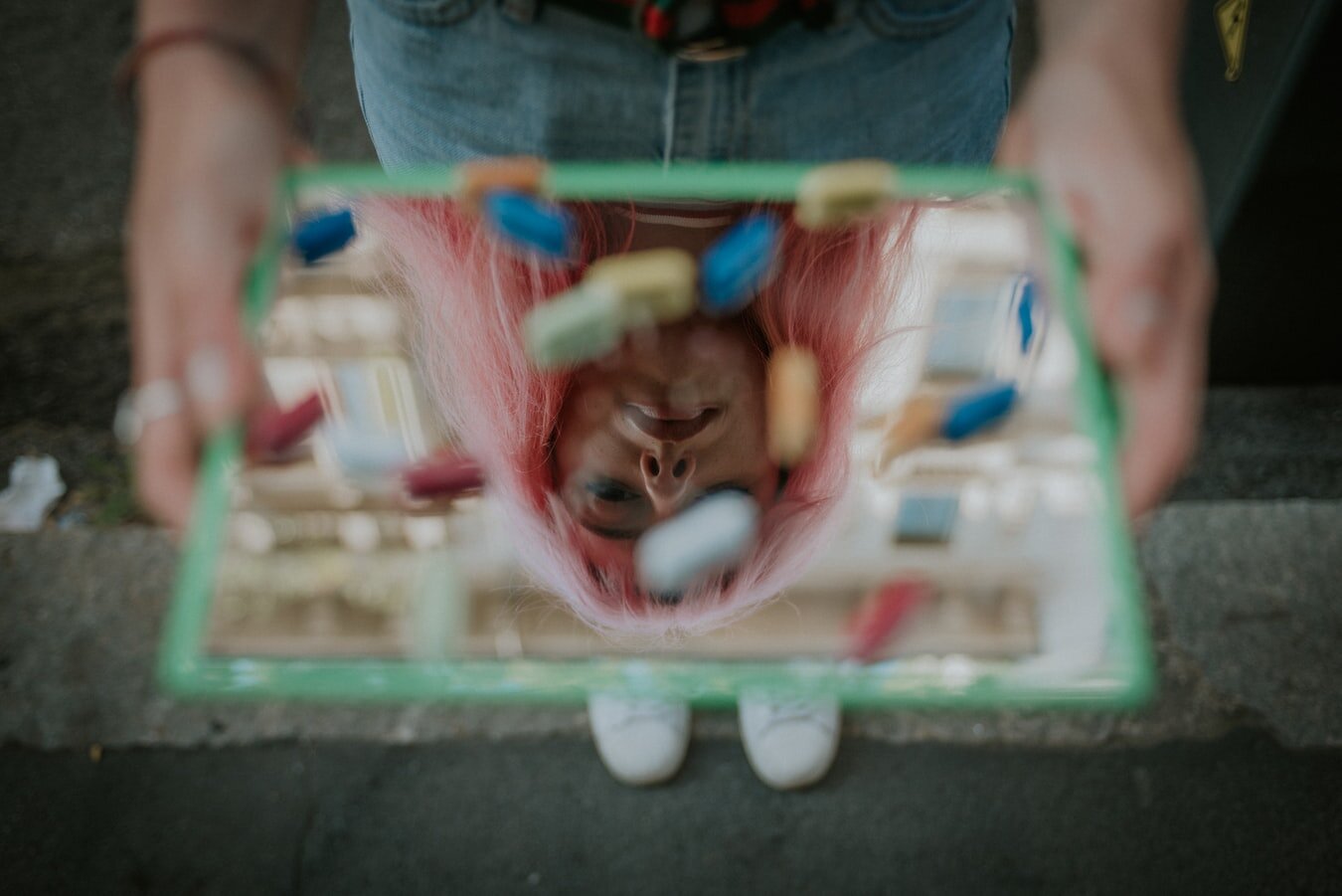Woman looking downward at her reflection in a mirror, with a pile of pills scattered on top of it.