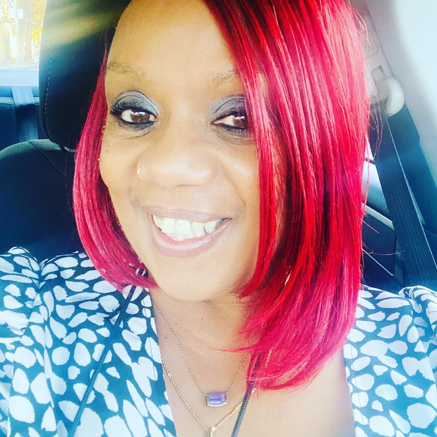 Story contributor Yolanda Morrissette has a bright-red, shoulder-length bob and is wearing a blue and white top, and is smiling at the camera from the driver's seat of her car
