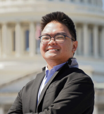 Aaron Facundo standing, arms folded, smiling, wearing a blazer, with the US Capitol building in the background.