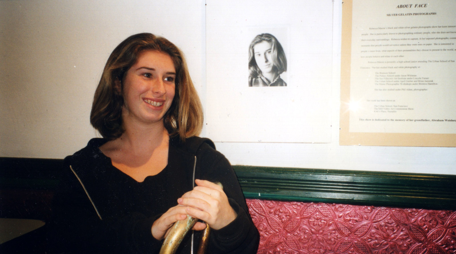 A young, smiling Becca sitting in front of her self-portrait at a photography show.