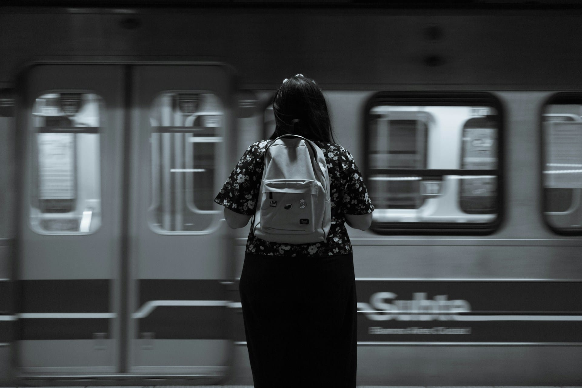 Black and white photo of a woman standing in front of a subway train passing by.