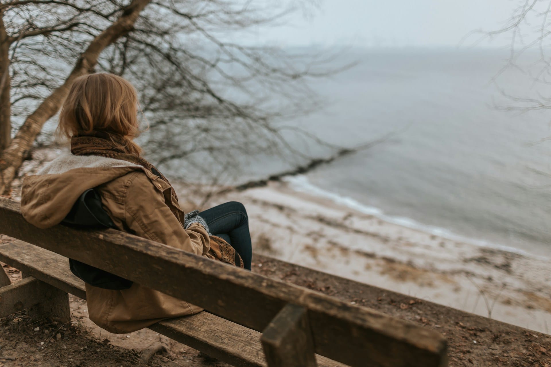 Woman sitting on brown wooden bench, looking out into the ocean.