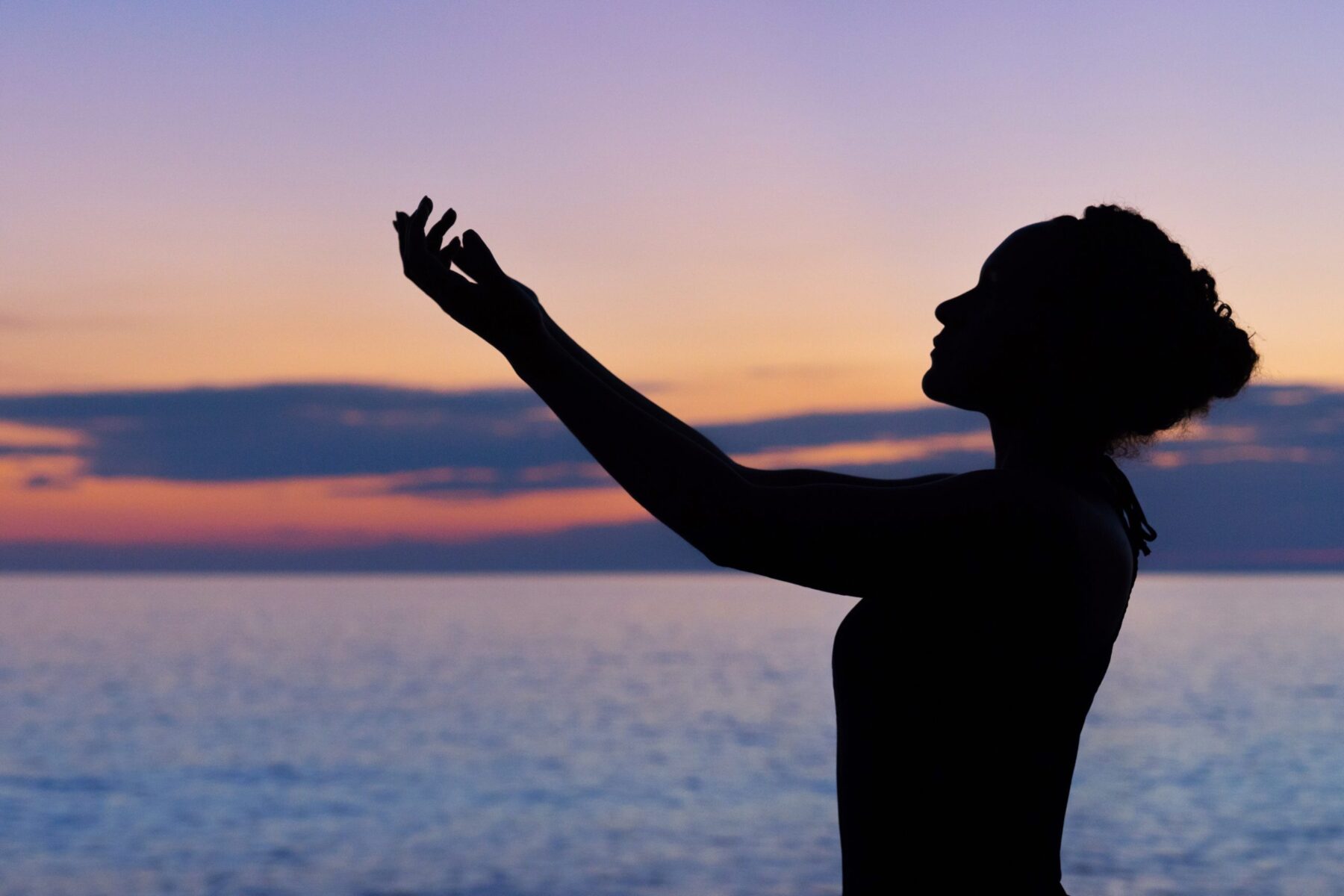 Silhouette of a woman's profile standing in front of a lake reaching her arms up to the sky.