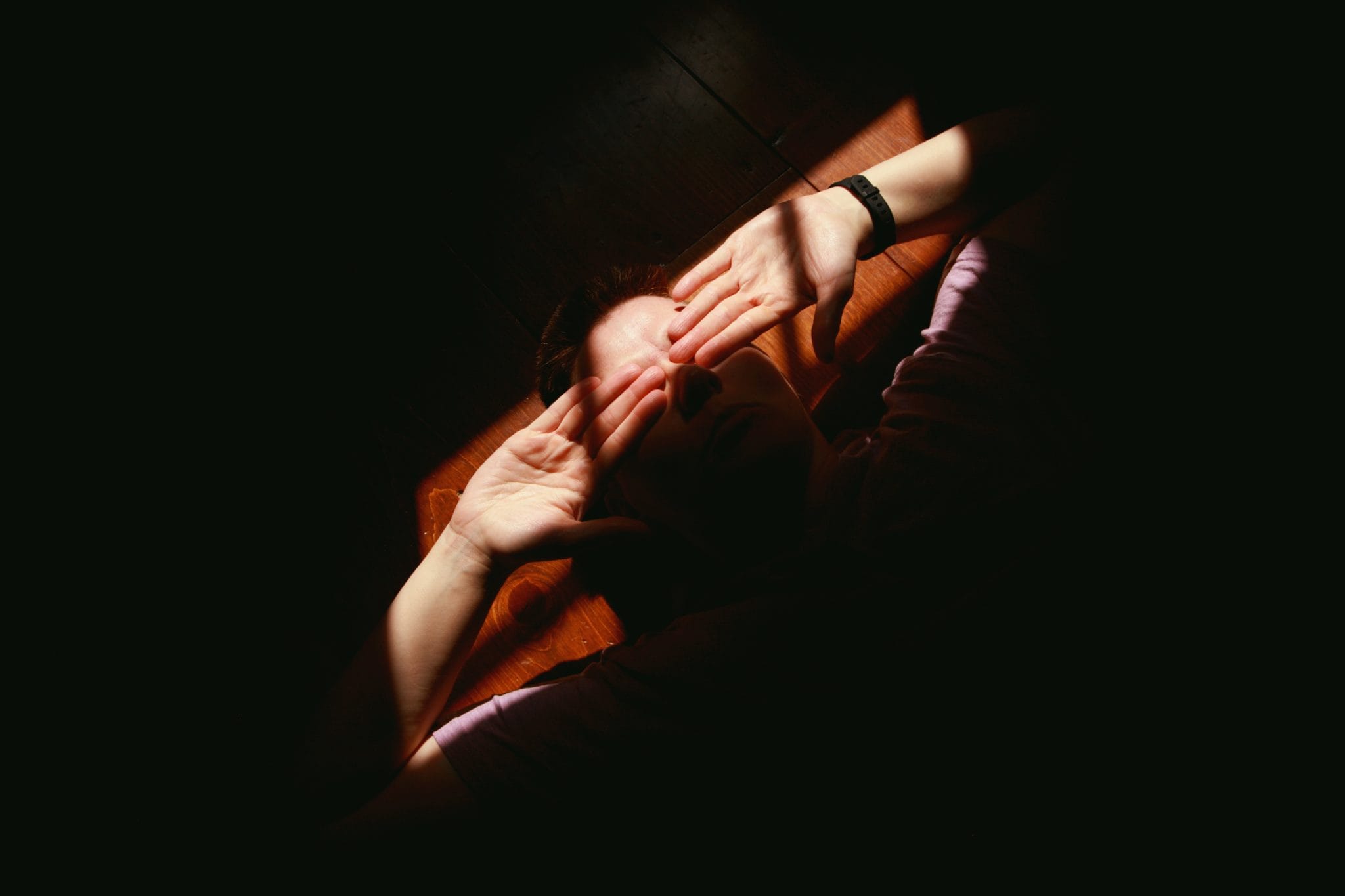 A person lying on the floor, with sunlight illuminating their hands held over their face.